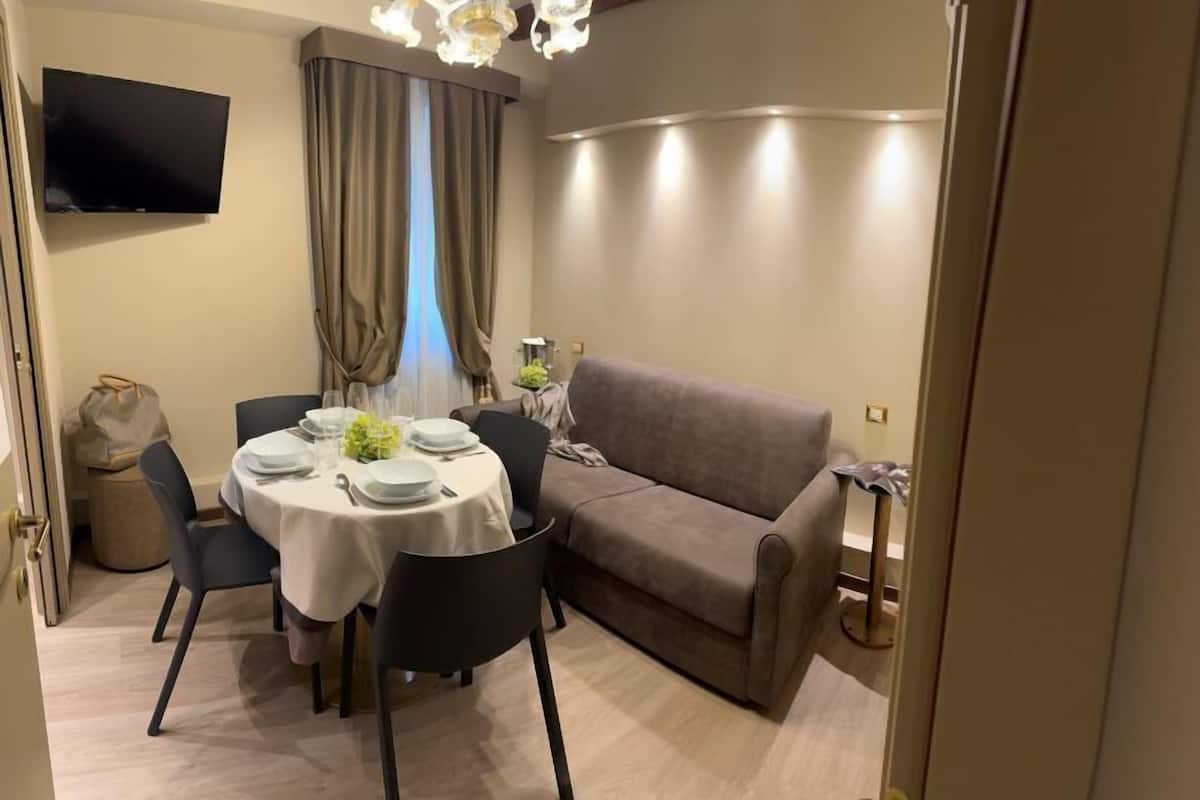 A dining area with a round table set for four, a sofa, a television mounted on the wall, and a window with curtains.