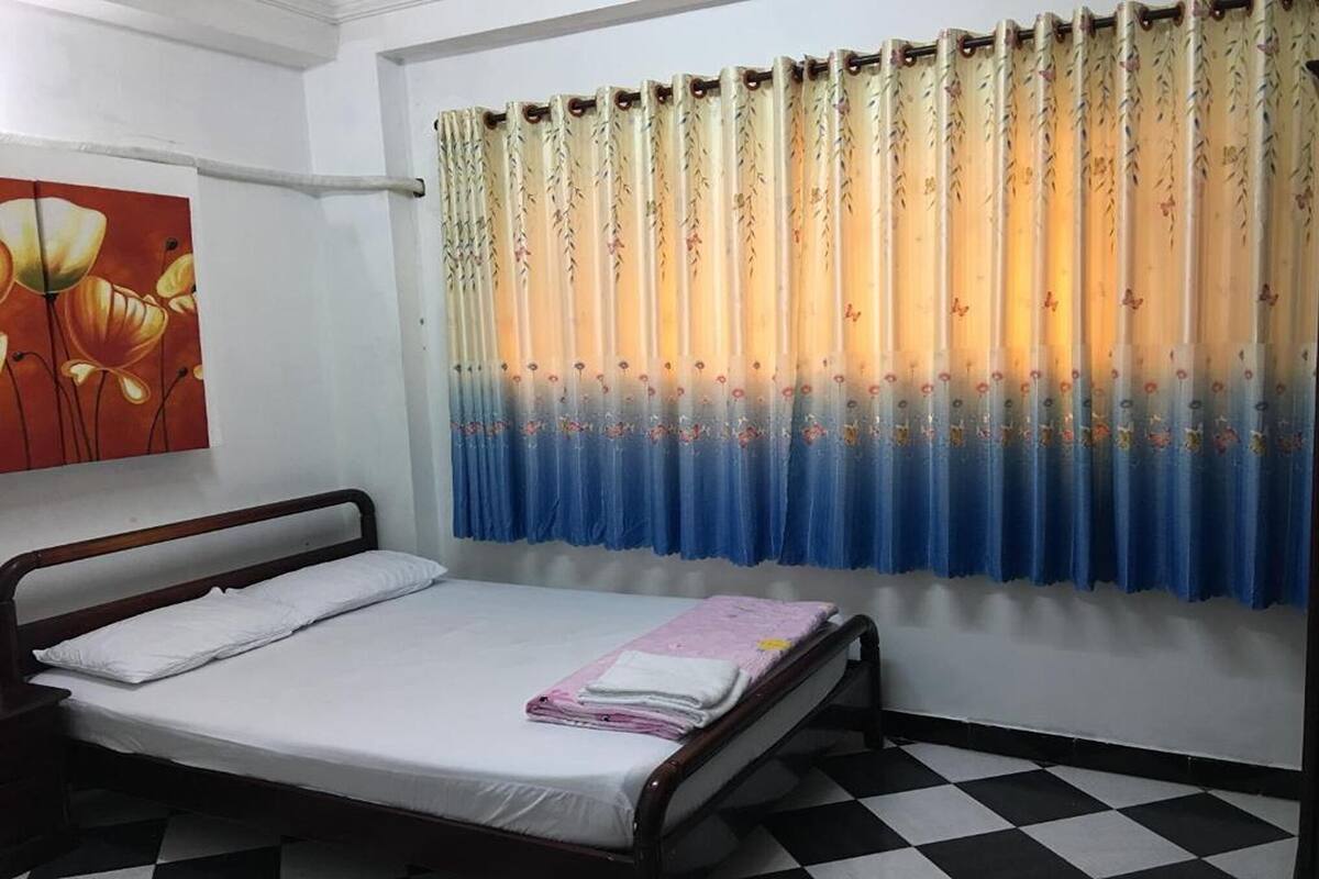 A single bed with a white mattress and a wooden frame, a blue curtain with floral patterns, and a checkered floor.