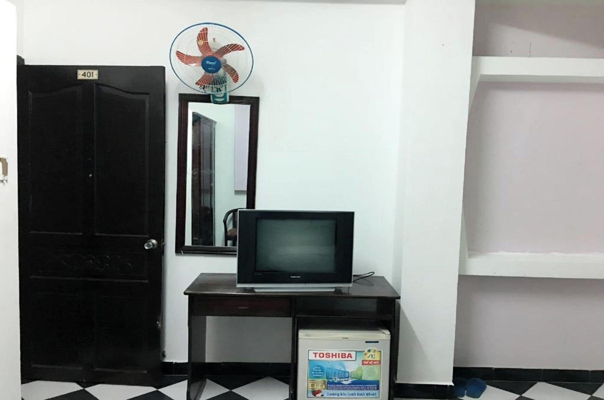Quarto com porta fechada, escrivaninha com televisão, espelho e refrigerador Toshiba.