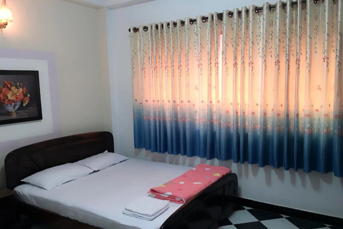 A bedroom with a bed, a nightstand, a checkered floor, and a window with floral curtains.