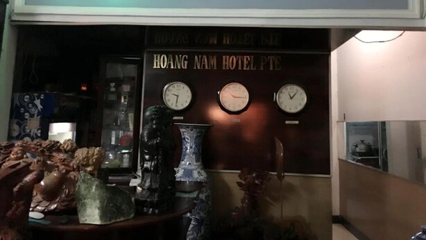 The exterior of Hoang Nam Hotel with a neon sign, three clocks on the wall, and decorative items on the counter. - Hoang Nam Hotel by Zuzu (Ho Chi Minh Municipality)