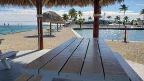 Un área frente a la playa con un banco de madera, palmeras, una estructura de techo de paja y una piscina.