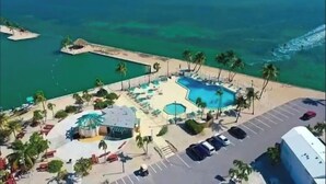 Vista aérea de un complejo turístico con una piscina grande, palmeras y un estacionamiento.