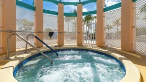 Un jacuzzi con una escalera de metal, rodeado por una cerca de celosía blanca y pilares color beige, bajo un cielo azul despejado.