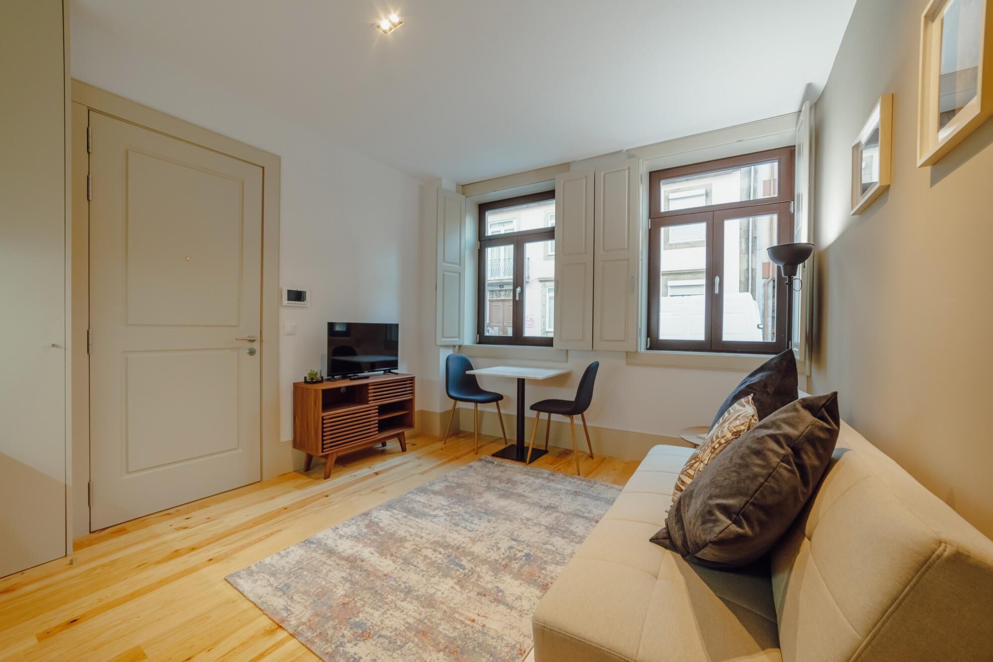 Un espacio habitable compacto con un sofá, un televisor sobre un soporte de madera, una mesita con dos sillas y una ventana con vistas a edificios.