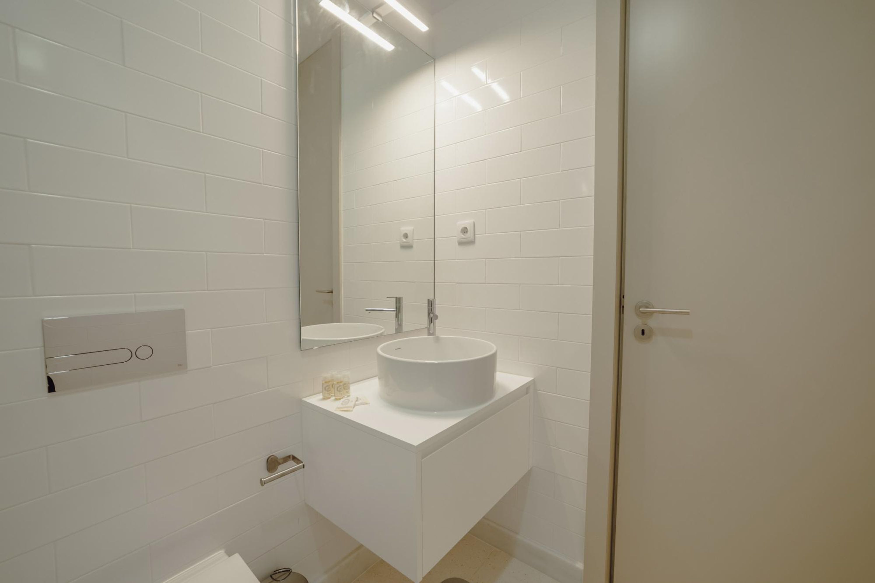 Un baño moderno con baldosas blancas, un espejo grande, dos lavamanos y un inodoro.