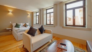 A modern hotel room with a bed, a sofa, a coffee table, and large windows offering views of the city.