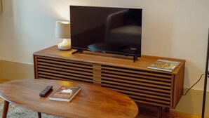A modern living room with a flat-screen TV on a wooden stand, a small table with a remote and a magazine, and a floor lamp.