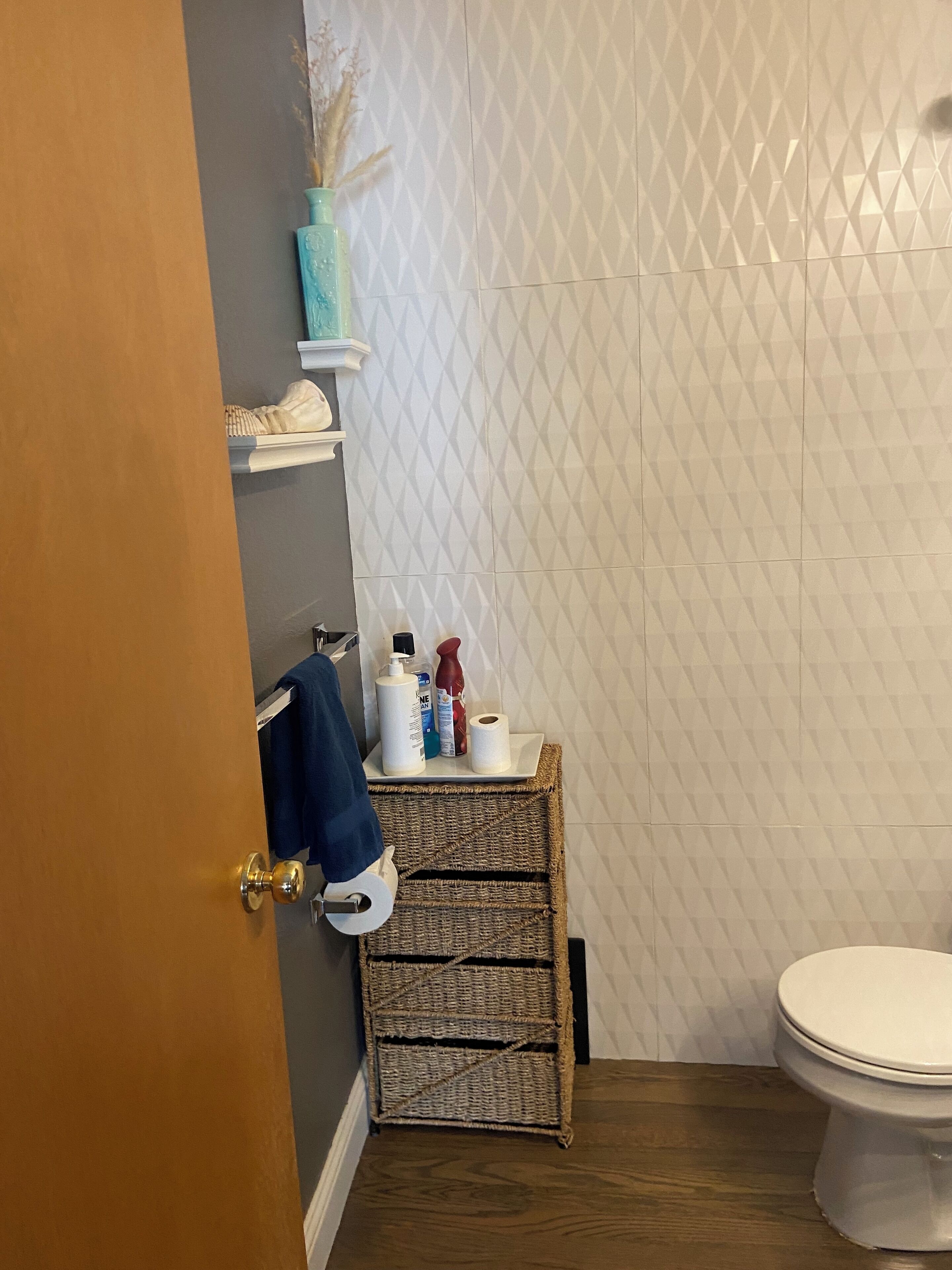 Un baño con inodoro blanco, suelo de madera, un perchero con toalla azul y un estante con artículos de aseo.