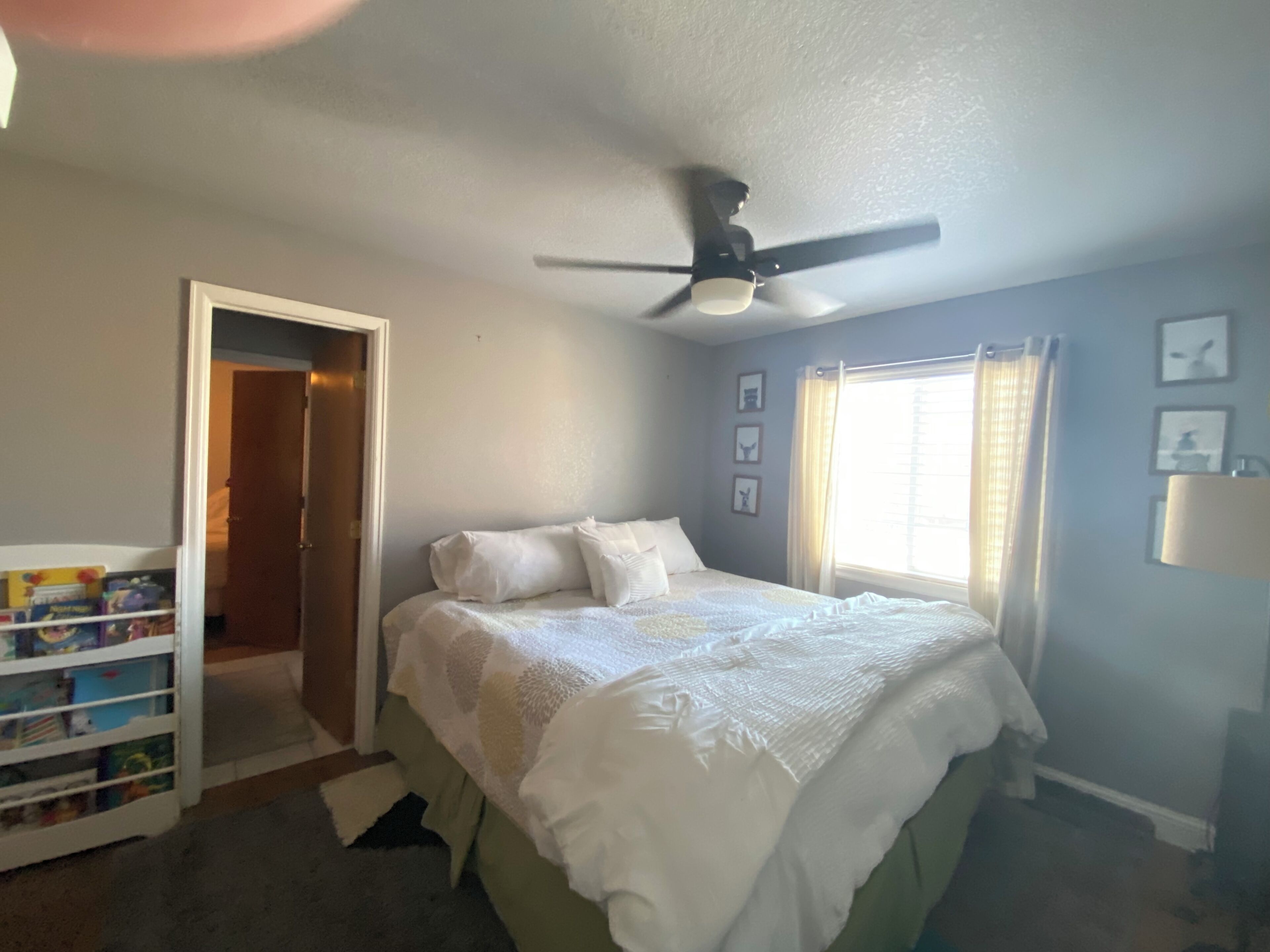 Un dormitorio con cama, ventilador de techo, ventana con cortinas y una puerta que da a otra habitación.