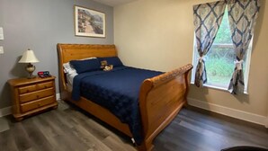 Un dormitorio con cama de madera, mesita de noche, lámpara, ventilador de techo, ventana con cortinas e imagen en un marco en la pared.