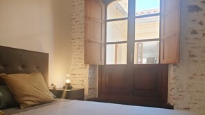 A bedroom with a large bed, a nightstand with a lamp, a window with wooden shutters, and a brick wall. - Picasso neighborhood. Beautiful flat in central 2B (Málaga)