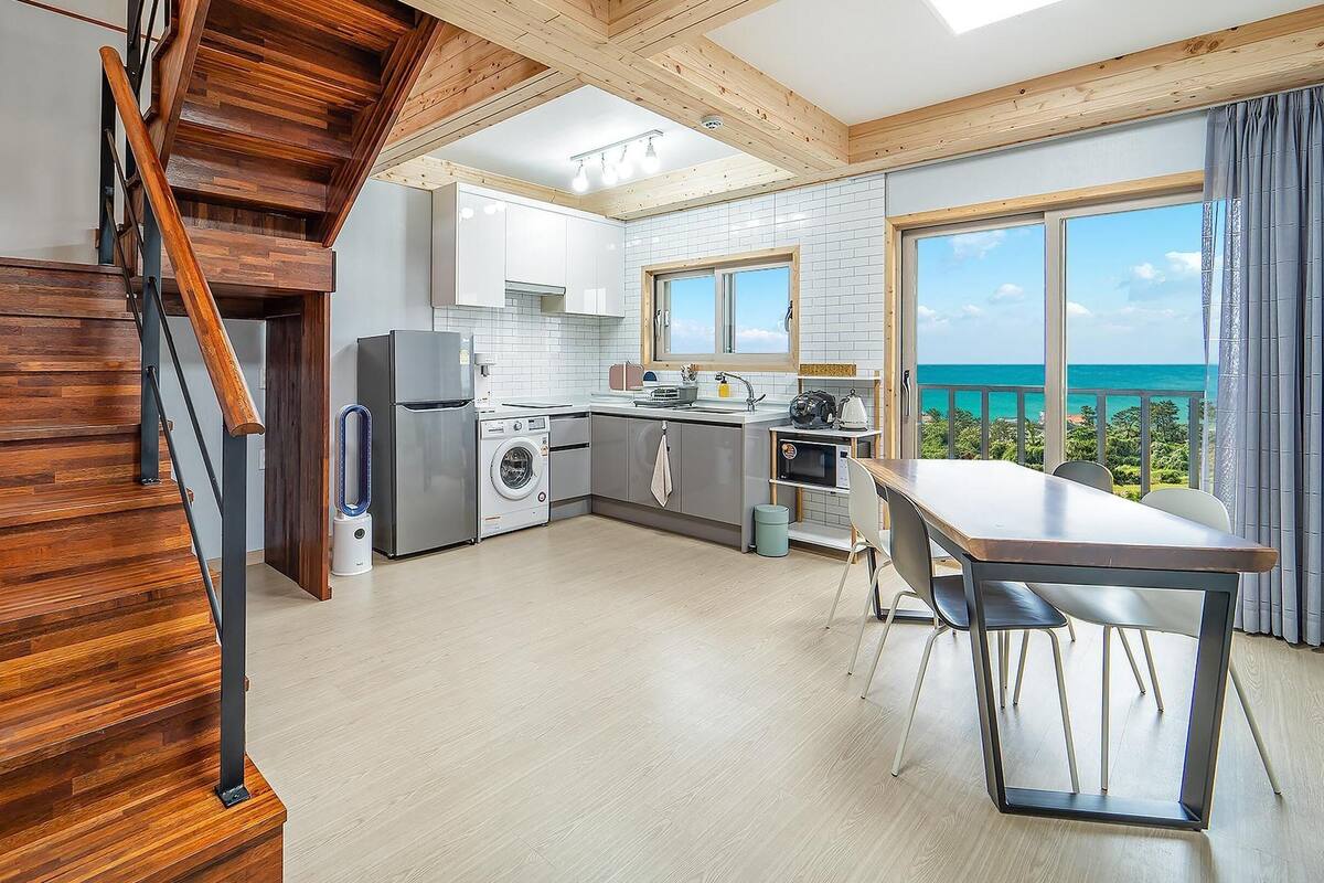 A modern kitchen with a view of the ocean, wooden staircase, and a dining area with a table.