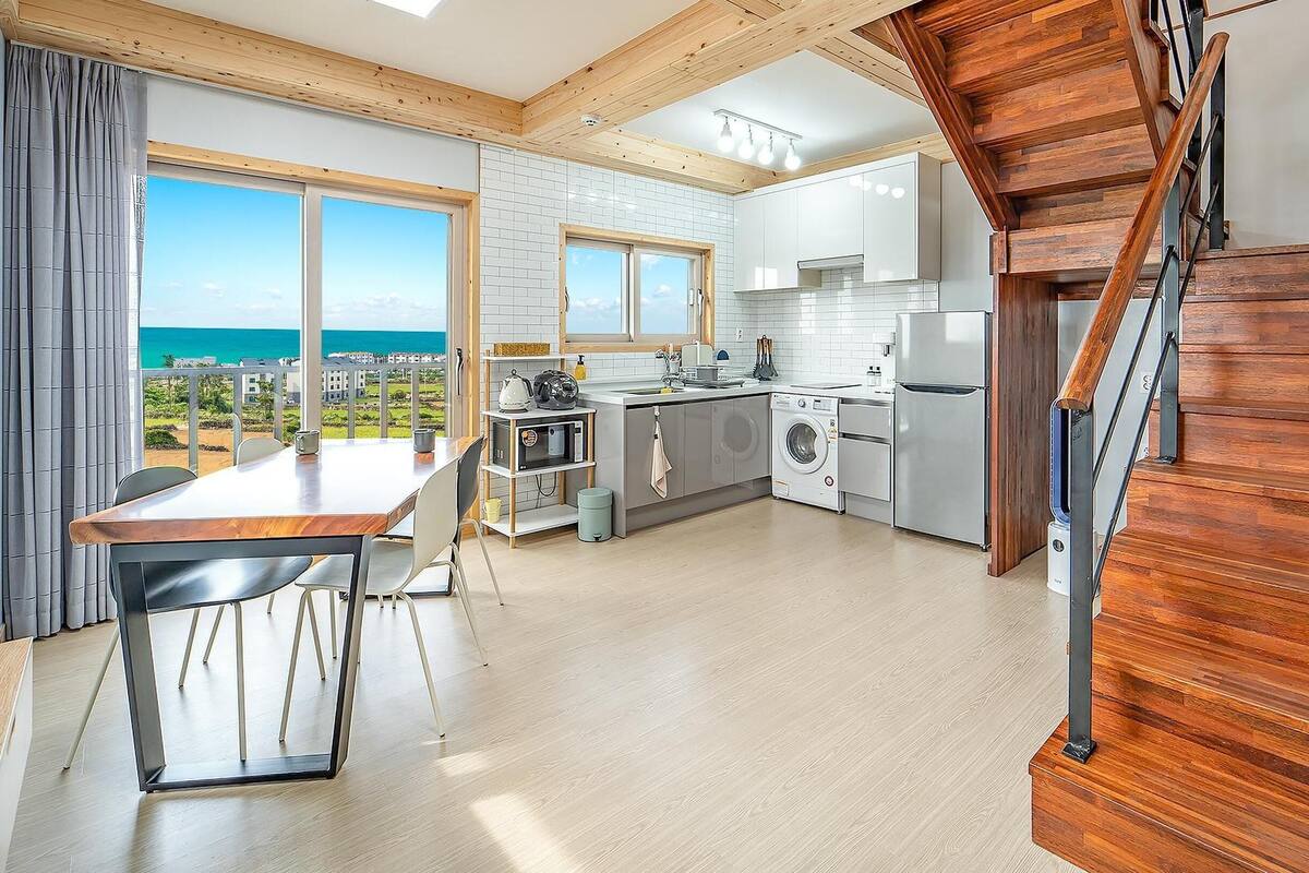 A modern kitchen with a view of the ocean, wooden beams, and a dining area with a table and chairs.