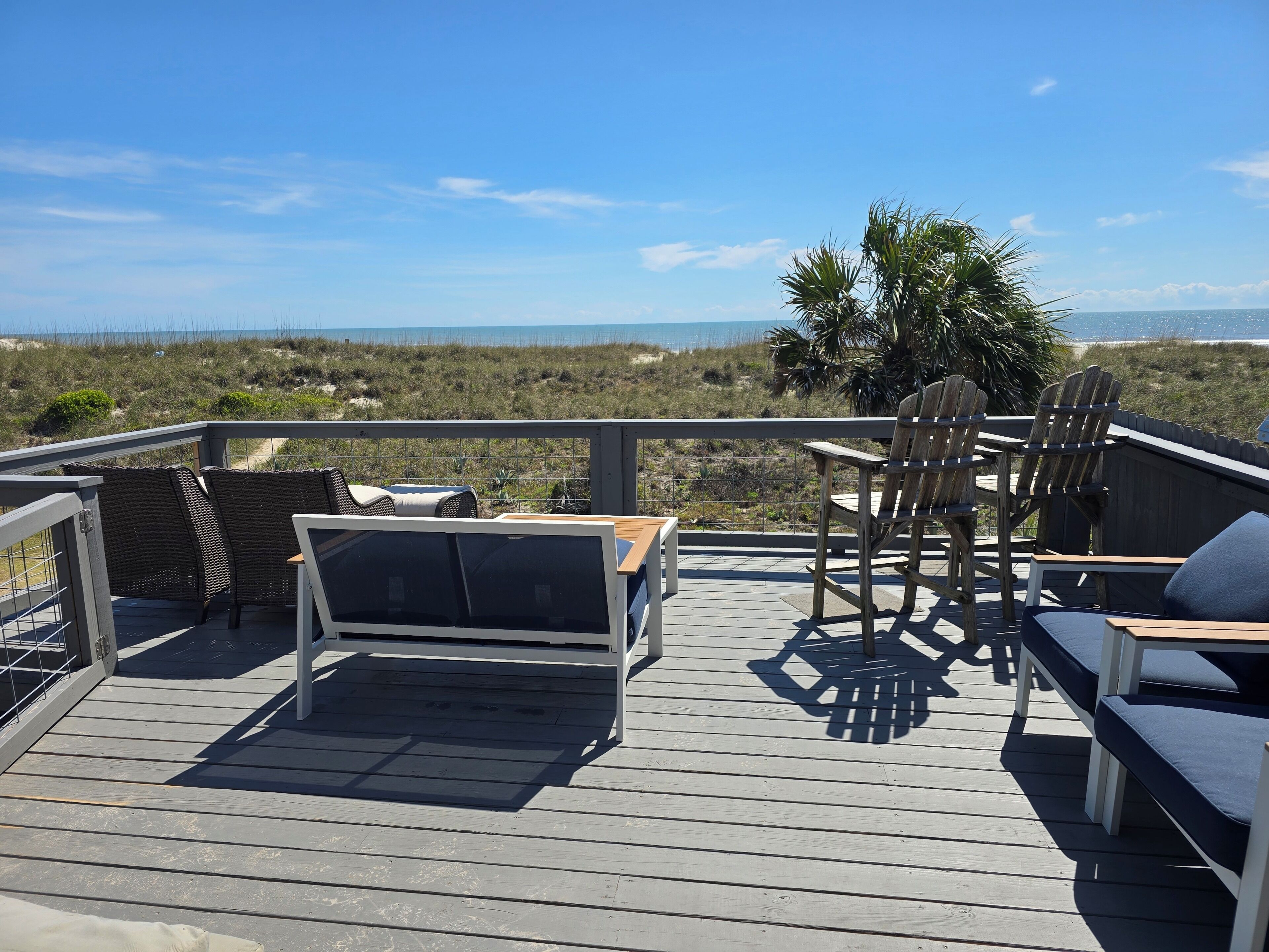 A wooden deck with outdoor furniture, including a sofa and chairs, overlooking a beach with dunes and a clear sky.