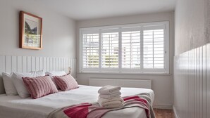 A neatly made bed with white and pink bedding, a window with blinds, and a framed picture on the wall. - Kirrary Townhouse 4 bedroom house VRBO (Dingle)