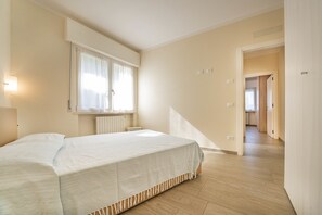 Une chambre à coucher avec un lit, une fenêtre avec des rideaux, un radiateur et un couloir menant à la cuisine.
