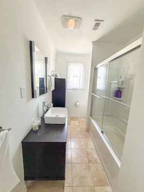 A bathroom with a dark countertop sink, a mirror, a window with white curtains, and a glass shower enclosure. - Pet friendly beach house w/ Hot Tub - Short 0.4 mile walk to beach! (Kill Devil Hills)