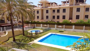 A swimming pool surrounded by a well-maintained lawn, palm trees, and a building with multiple windows and balconies. - A&N Valle Niza (Benajarafe)