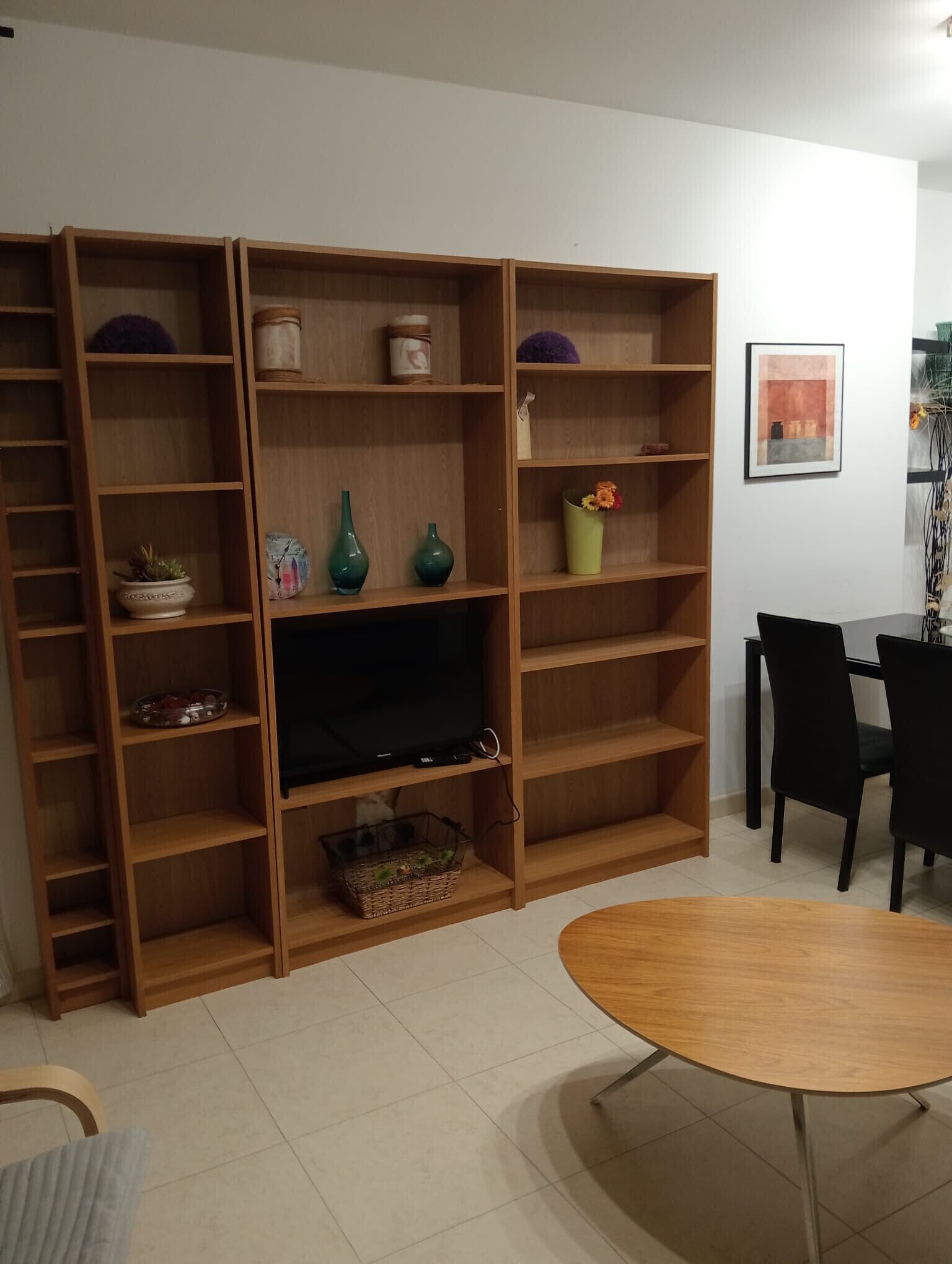 Uma sala de estar com TV em um móvel, prateleiras de madeira com objetos decorativos, uma mesa de jantar com cadeiras e um quadro na parede.