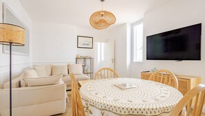 A living room with a sofa, a round dining table with chairs, a television mounted on the wall, and a wicker lamp.