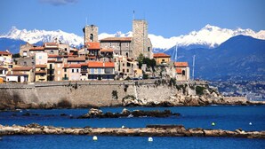 A coastal town with historic stone buildings, a prominent tower, and a backdrop of snow-capped mountains.