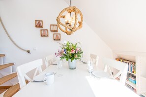 Zona pranzo con tavolo bianco apparecchiata per sei persone, circondata da sedie bianche. Un lampadario intrecciato pende sopra, e sul tavolo c'è un vaso con fiori.