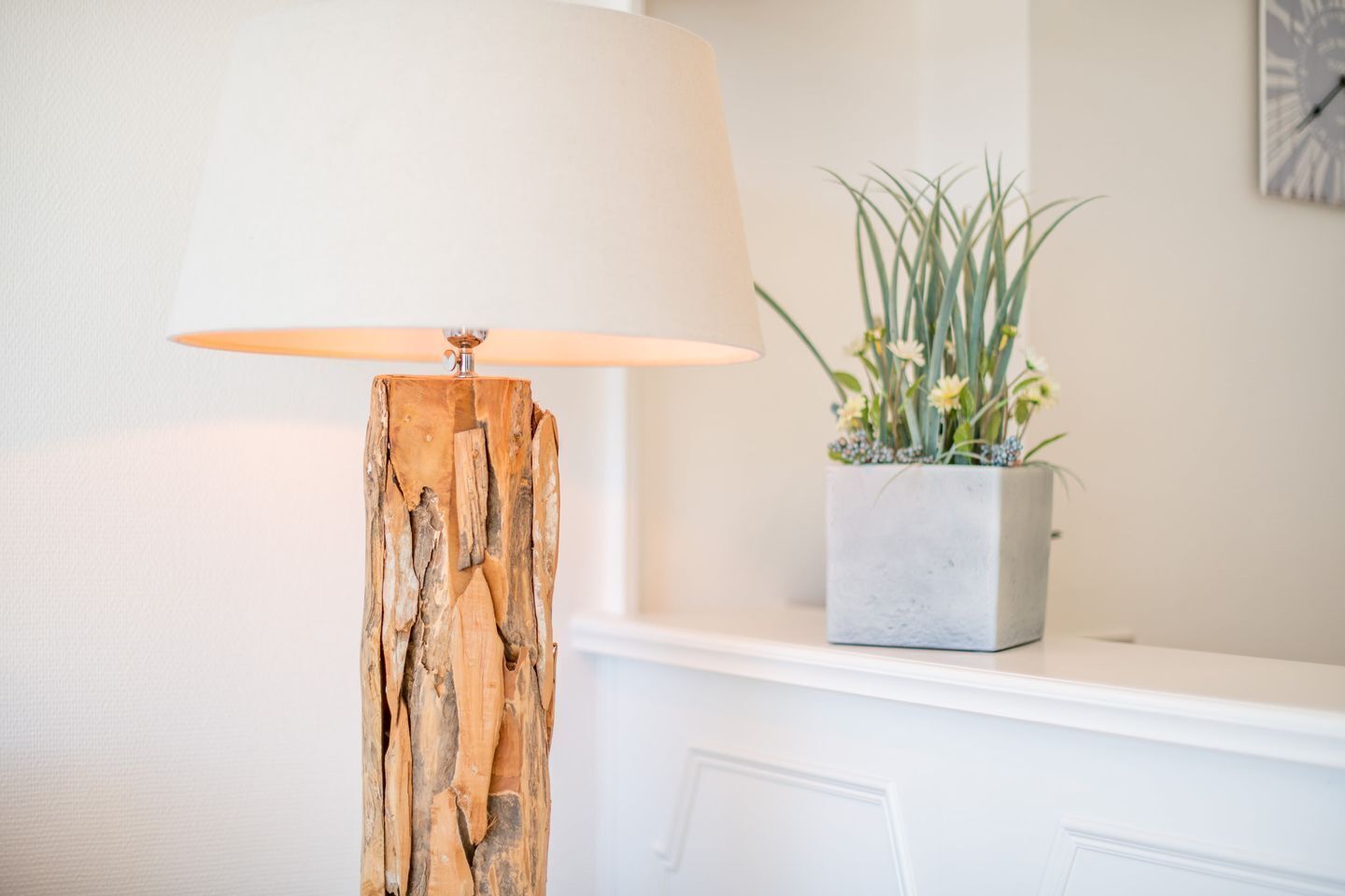 A floor lamp with a wooden base and a white lampshade, a potted plant with white flowers, and a white wall with a clock.
