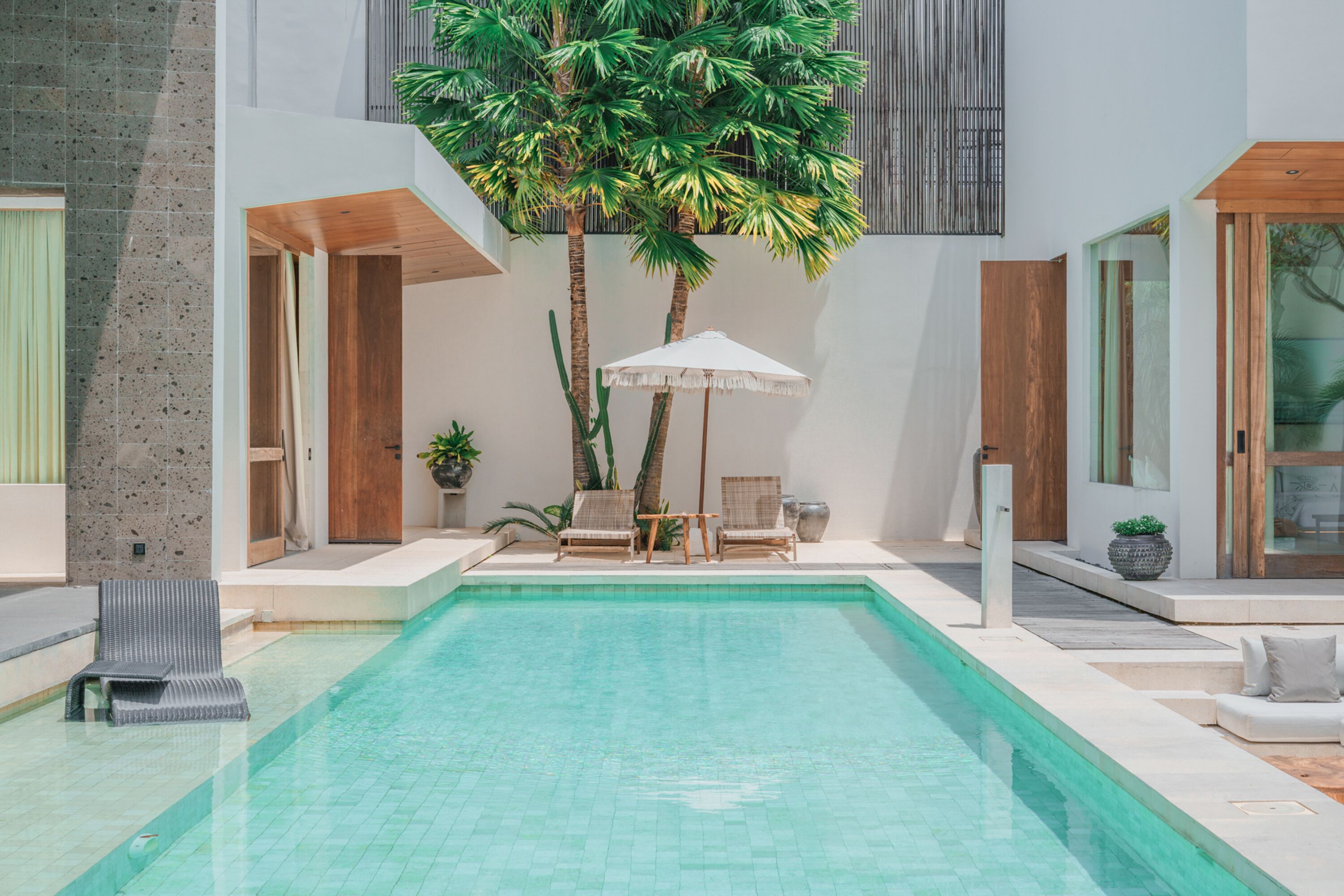 Un área de piscina moderna con una piscina cristalina, sillones de descanso y un gran árbol.