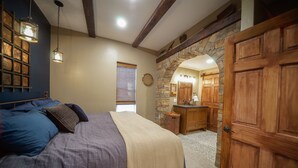 Une chambre à coucher avec un lit, une porte en bois, une arche en pierre et une salle de bain visible à travers l’arche.