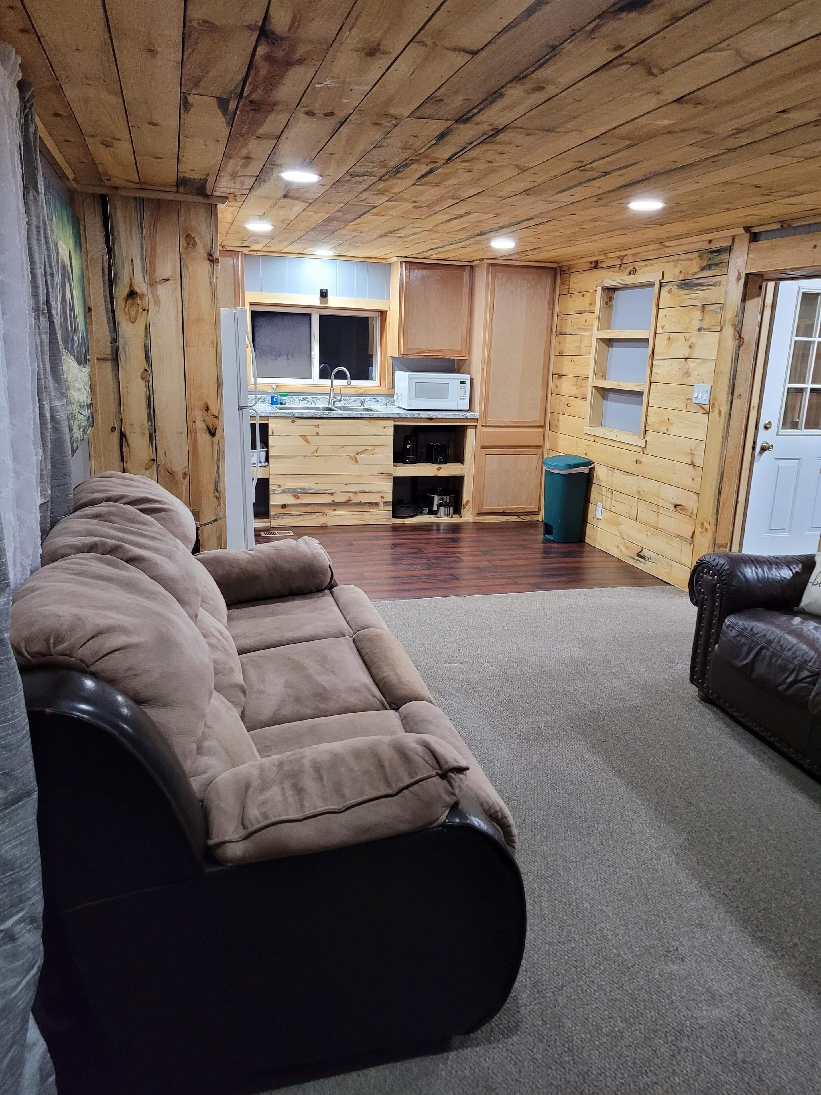 A cozy living room with a brown sofa, a wooden ceiling, and a kitchen area with wooden cabinets and a microwave.