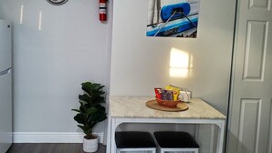 Une petite cuisine avec un plan de travail blanc, deux chaises noires, une plante en pot, une horloge murale, un extincteur et un tableau représentant un bateau.