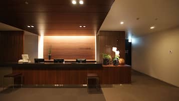 A reception desk with a wooden finish, a computer, and a telephone. There are potted plants and a small seating area with a bench.