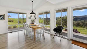 A spacious dining area with a wooden table and white chairs, large windows with a view of a green landscape, and a black chair. - Manandel - Rural Retreat (Bonnie Doon)