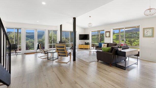 A spacious living room with a large sofa, a coffee table, and a TV. There are multiple windows offering a view of the outdoors. - Manandel - Rural Retreat (Bonnie Doon)