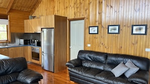 Un interior de cabaña acogedor, con detalles de madera, una kitchenette, un sofá de cuero negro y una mesa de comedor.