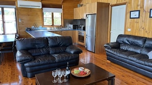 Interior moderno de cabaña con paredes de madera, un área de cocina, dos sofás de cuero, y una mesa de comedor con copas de vino y una bandeja de frutas.