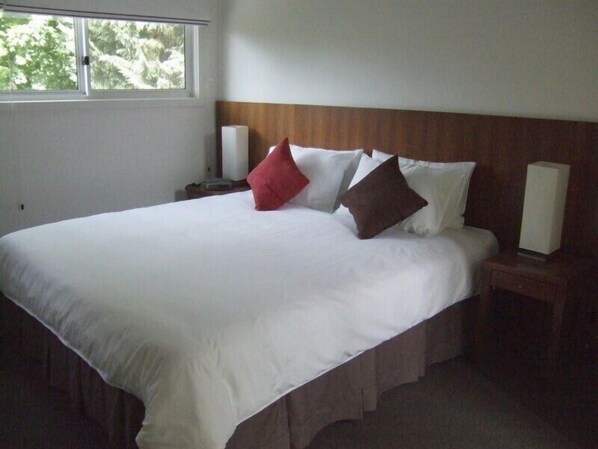 A hotel room with a large bed, white linens, a wooden headboard, two bedside tables with lamps, and a window with a view of trees.