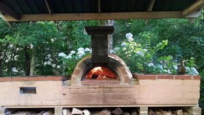 A brick-built outdoor pizza oven with a fire burning inside, surrounded by stacked firewood and greenery. - Goulburn River Cottage - River Front (Jamieson)