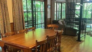 A wooden dining table with chairs, large windows with curtains, a staircase, and a sofa in a room with wooden floors. - Goulburn River Cottage - River Front (Jamieson)