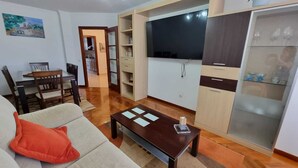 Una sala de estar con sofá, mesa de centro y televisor. Hay una zona de cocina con armarios y refrigerador.