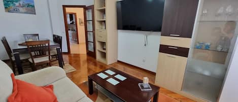 A living room with a sofa, coffee table, and a TV. There is a kitchen area with cabinets and a refrigerator.
