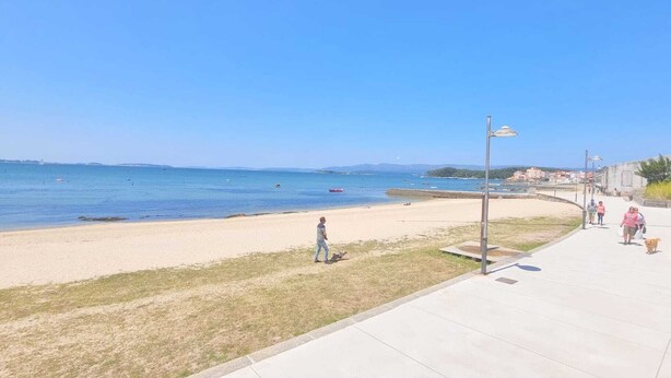 Una playa con un paseo marítimo pavimentado, una zona ajardinada y un cielo azul despejado.