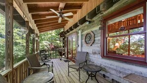 Una terraza de madera con mobiliario exterior, un ventilador de techo y un ventanal con vistas a árboles.
