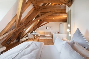 Une chambre de style « loft », avec des poutres en bois apparentes, un lit recouvert de linge de maison blanc, un petit bureau et un canapé à l’arrière-plan.