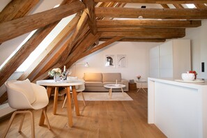 Une pièce mansardée avec des poutres apparentes, un canapé, une table à manger avec des chaises, une petite table ronde et un comptoir blanc.