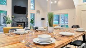 Una mesa de comedor preparada con platos blancos, tazones y vasos transparentes, rodeada de sillas, en una habitación con chimenea, ventanas amplias y un televisor montado en la pared.
