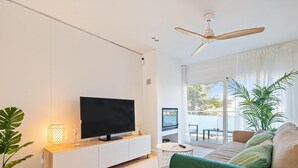 Una sala de estar moderna con un televisor de pantalla plana sobre una consola blanca, un ventilador de techo, un ventanal con cortinas y un sofá verde.