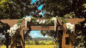 A wooden arbor with white flowers, a bench, and a grassy field in the background. - Lake Road Cabin C — Million Dollar Ozark View! (Ironton)