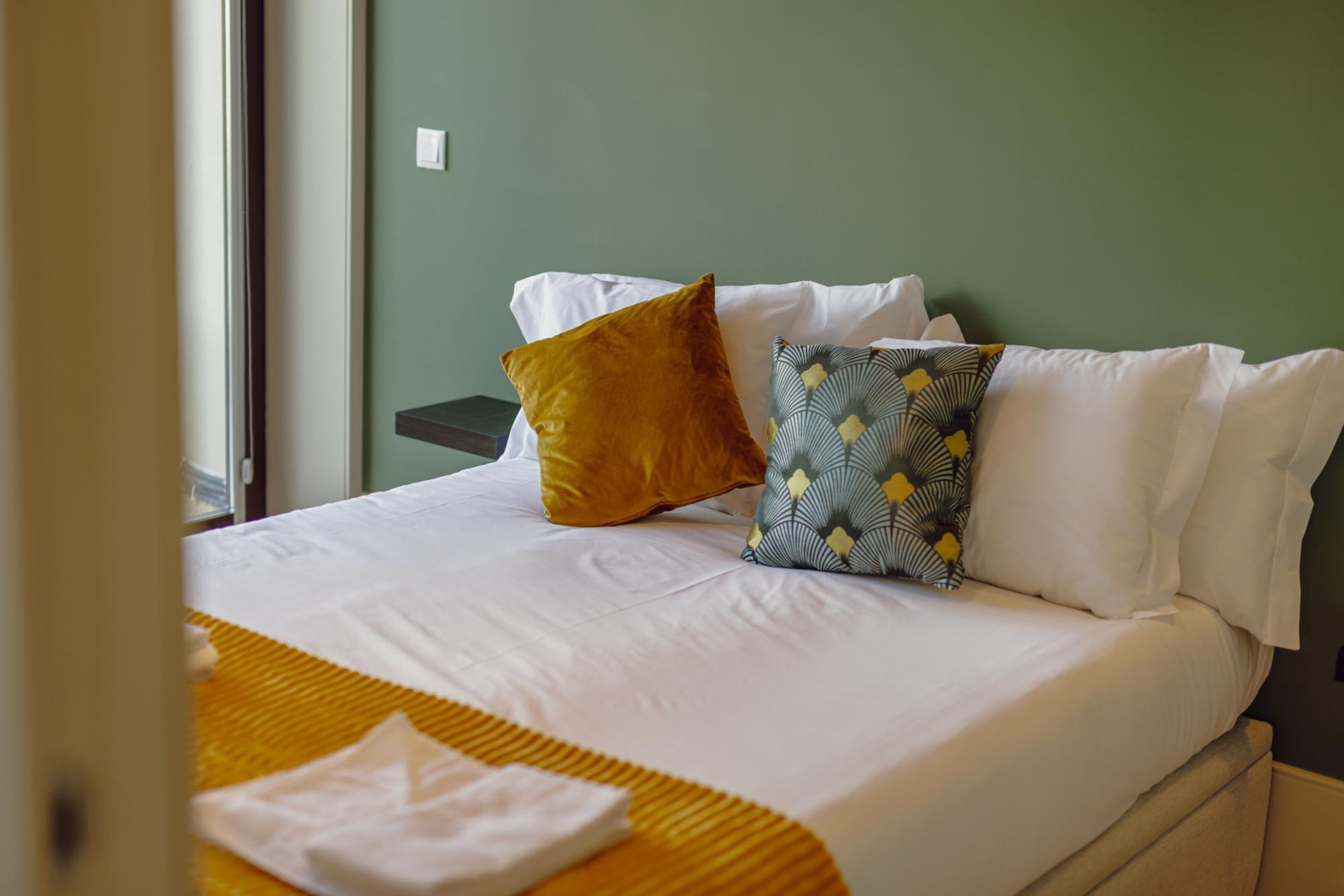 Un lit bien fait, avec du linge de lit blanc, un plaid de couleur moutarde et des coussins à motifs.